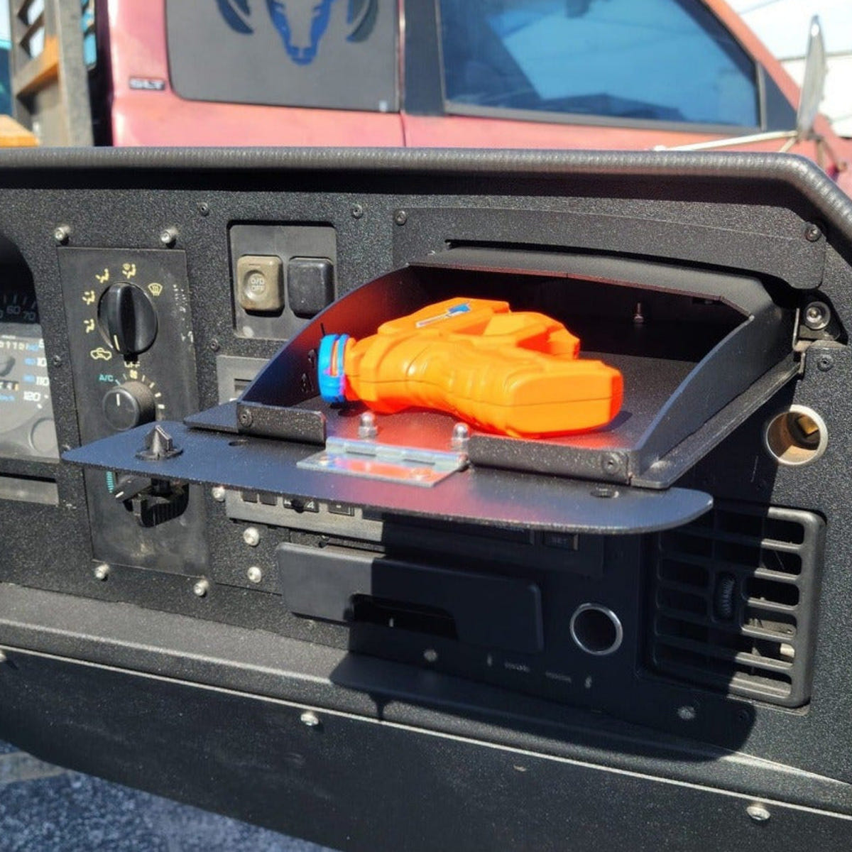 A black metal truck bed accessory shelf mounted on the side of a red pickup truck, holding an orange item. The photo is taken outdoors in daylight, with the truck bed and gravel ground visible.
